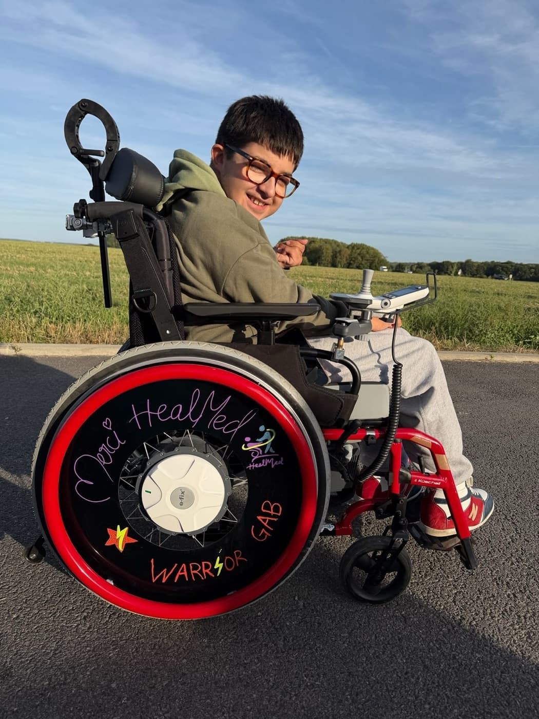 Un jeune garçon souriant dans son fauteuil roulant électrique, « Merci HealMed » écrit sur la roue